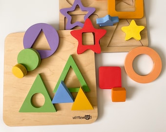 Op de afbeelding: Een houten puzzelset met kleurrijke vormen, waaronder een ster, een cirkel, een vierkant en een driehoek. De vormen zijn uit de houten planken geknipt en kunnen op de planken worden geplaatst. De puzzelset is gemaakt door het bedrijf "Wittlewood".