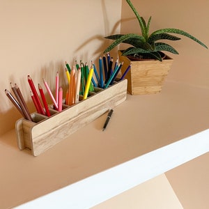 May include: A wooden pencil holder with six compartments, filled with colored pencils and a black pen. The holder is sitting on a shelf with a potted succulent plant in the background.