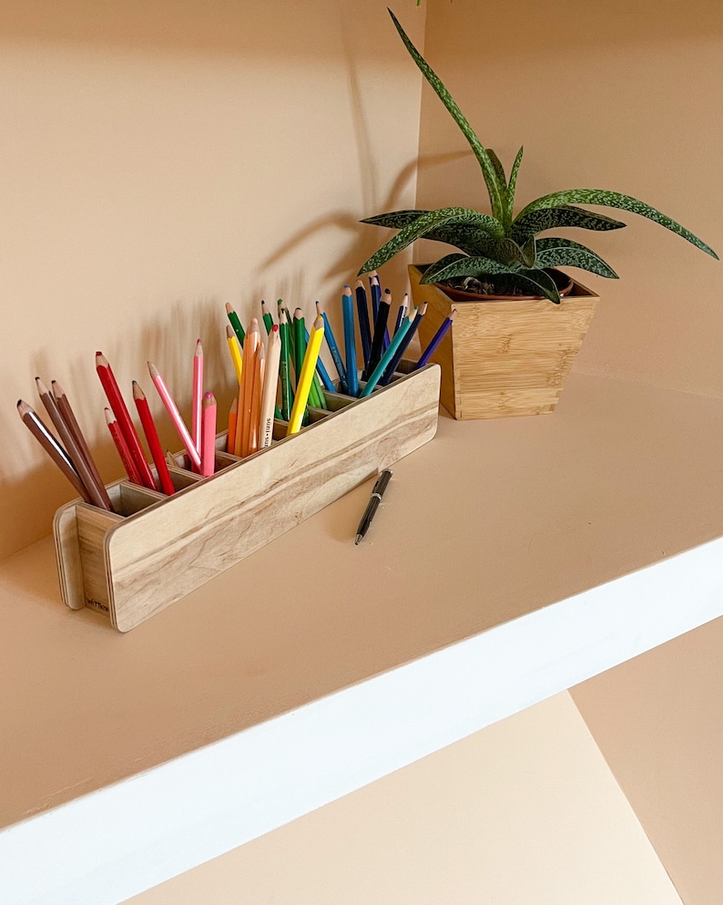 May include: A wooden pencil holder with six compartments, filled with colored pencils and a black pen. The holder is sitting on a shelf with a potted succulent plant in the background.