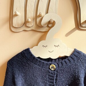 May include: A wooden cloud-shaped hanger with a smiling face and closed eyes. The hanger is hanging on a wooden rainbow wall decor. A black knitted cardigan with buttons is hanging on the hanger.