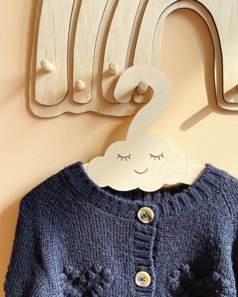 May include: A wooden cloud-shaped hanger with a smiling face and closed eyes. The hanger is hanging on a wooden rainbow wall decor. A black knitted cardigan with buttons is hanging on the hanger.