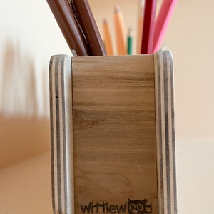May include: A wooden pencil holder with a light wood finish and white stripes on the sides. The holder has the text "wittlewood" and a small logo of a bird on the front.