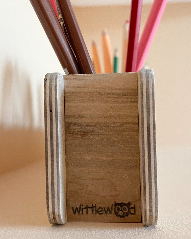 May include: A wooden pencil holder with a light wood finish and white stripes on the sides. The holder has the text "wittlewood" and a small logo of a bird on the front.