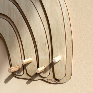 May include: A wooden rainbow wall hanger with four pegs. The rainbow is made of multiple layers of wood and has a natural wood finish. The pegs are made of wood and are attached to the bottom of the rainbow. The rainbow has the text "Wittlewood" printed on the bottom.