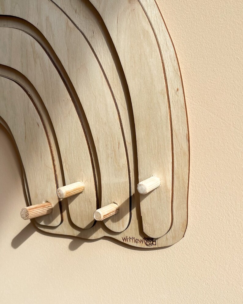 May include: A wooden rainbow wall hanger with four pegs. The rainbow is made of multiple layers of wood and has a natural wood finish. The pegs are made of wood and are attached to the bottom of the rainbow. The rainbow has the text "Wittlewood" printed on the bottom.