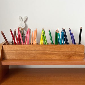 May include: A wooden pencil holder with multiple compartments, filled with colored pencils in a rainbow pattern. The holder is on a wooden shelf.