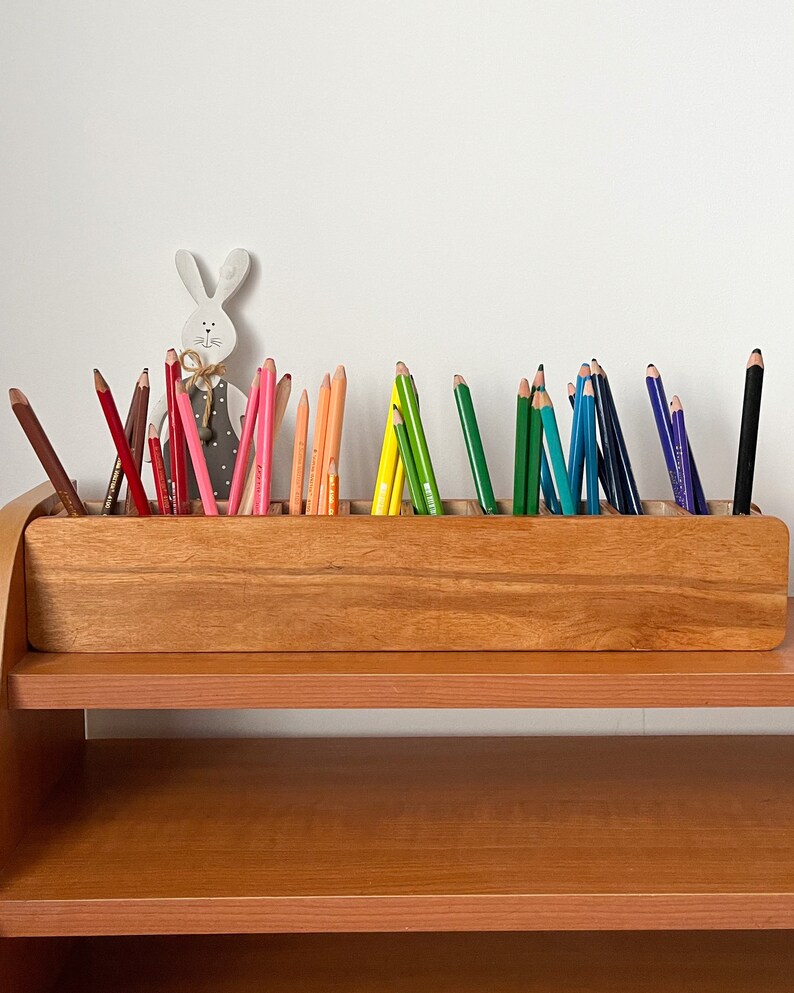 May include: A wooden pencil holder with multiple compartments, filled with colored pencils in a rainbow pattern. The holder is on a wooden shelf.