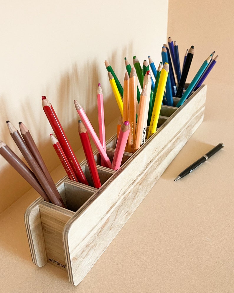 May include: A wooden pencil holder with six compartments, filled with colored pencils. The holder is made of light wood with a natural grain pattern. The text "WittleOwl" is visible on the bottom of the holder.