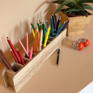 May include: A wooden pencil holder with six compartments, filled with colored pencils and black pens. The holder is made of light wood with a natural finish.