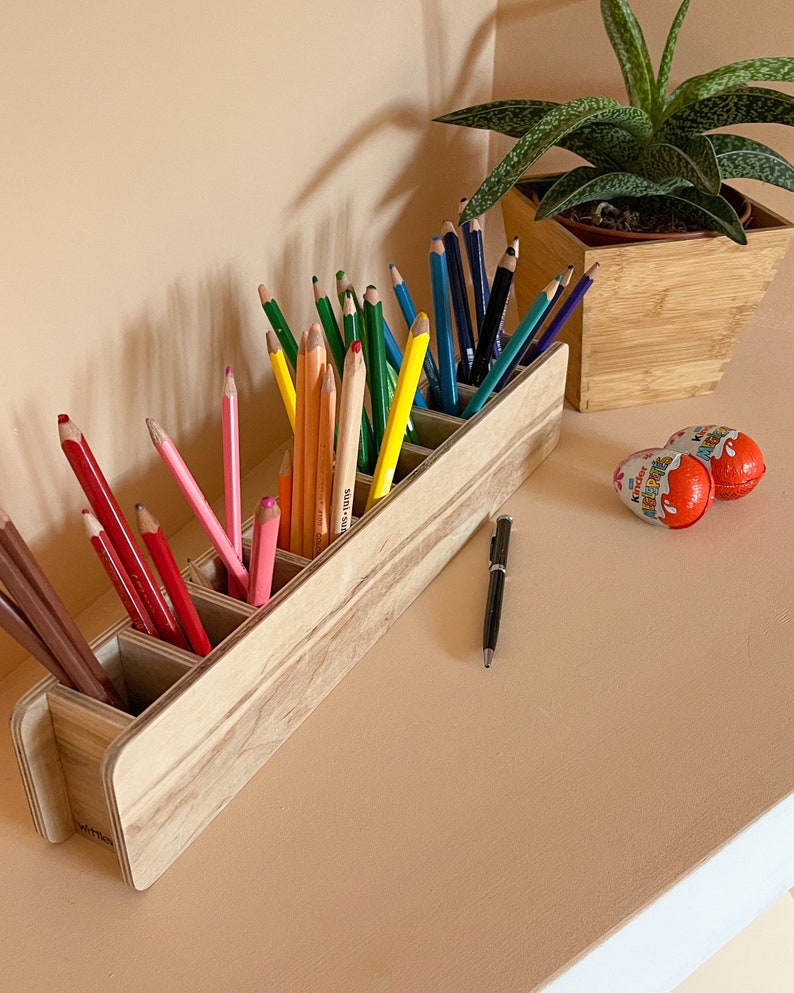 May include: A wooden pencil holder with six compartments, filled with colored pencils and black pens. The holder is made of light wood with a natural finish.