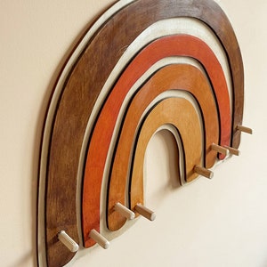 May include: A wooden rainbow wall decor with five layers of wood in shades of brown, orange, and white. The rainbow has eight wooden pegs for hanging items.