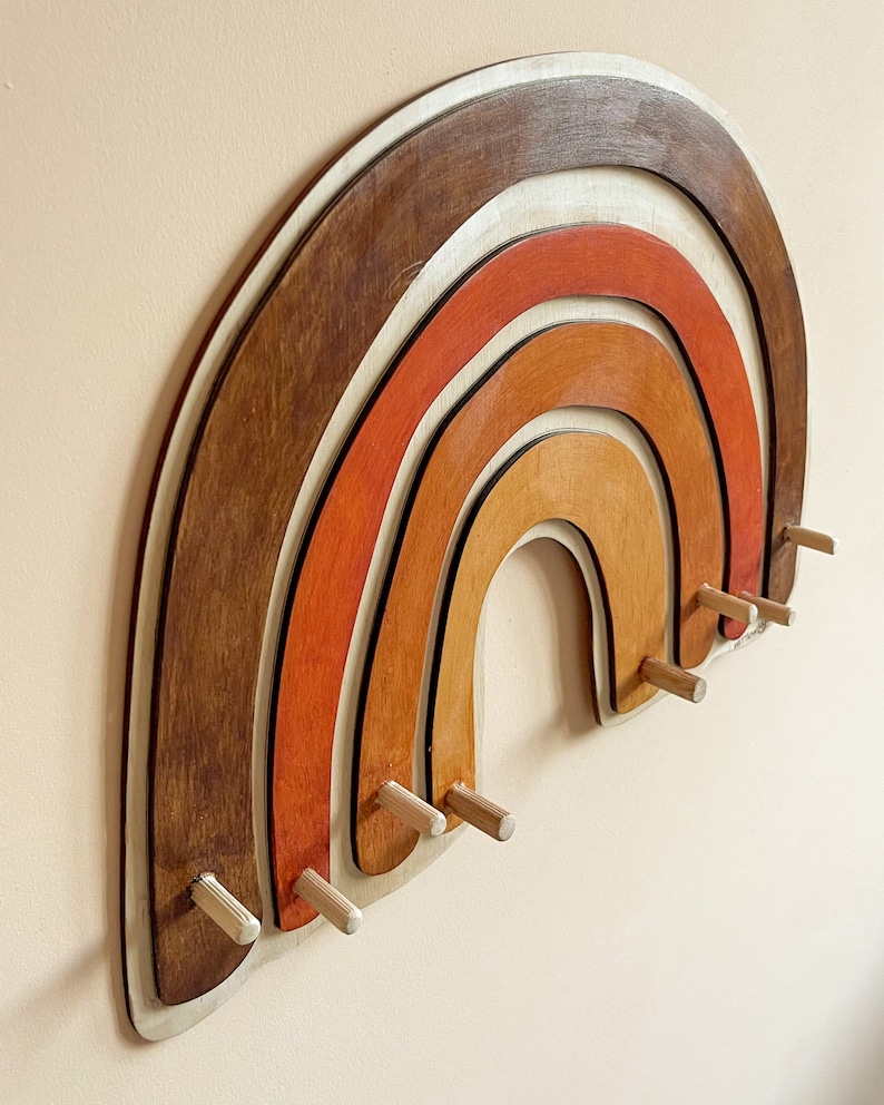 May include: A wooden rainbow wall decor with five layers of wood in shades of brown, orange, and white. The rainbow has eight wooden pegs for hanging items.