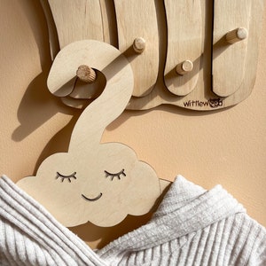 May include: A wooden cloud-shaped hanger with a smiling face and closed eyes. The hanger is made of light wood and has a natural finish. The hanger is hanging on a wooden wall rack with the text "Wittlewood" on it.