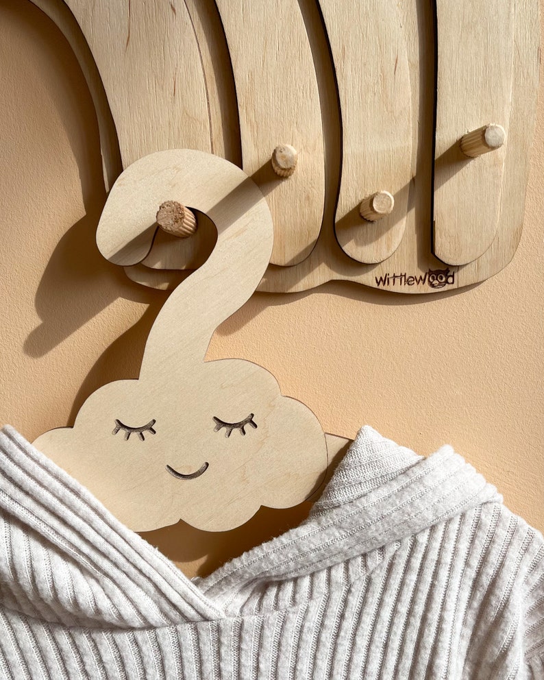 May include: A wooden cloud-shaped hanger with a smiling face and closed eyes. The hanger is made of light wood and has a natural finish. The hanger is hanging on a wooden wall rack with the text "Wittlewood" on it.