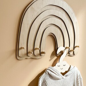 May include: Wooden rainbow wall hanger with five rounded arches and six wooden pegs. A white wooden cloud-shaped hanger with a smiling face is hanging from one of the pegs.
