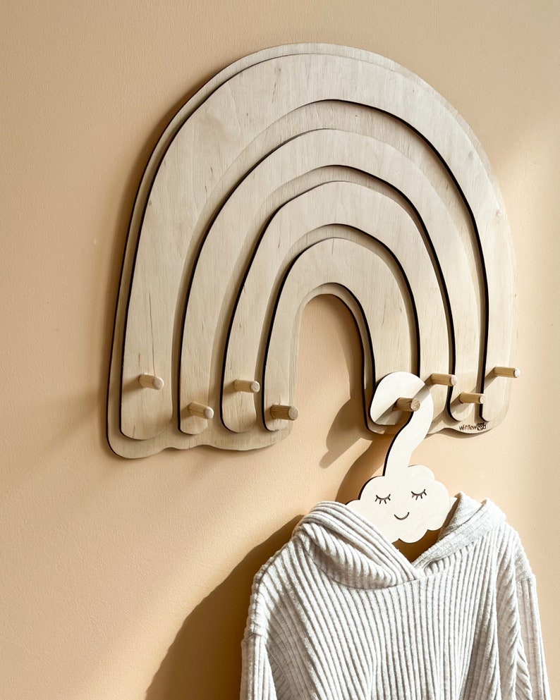 May include: Wooden rainbow wall hanger with five rounded arches and six wooden pegs. A white wooden cloud-shaped hanger with a smiling face is hanging from one of the pegs.