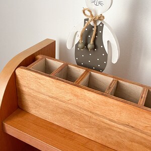 May include: A wooden desk organizer with a shelf and a compartment with multiple slots. The organizer is made of light brown wood and has a rounded top. A white wooden bunny figurine with a brown polka dot dress and a brown bow is standing on top of the organizer.