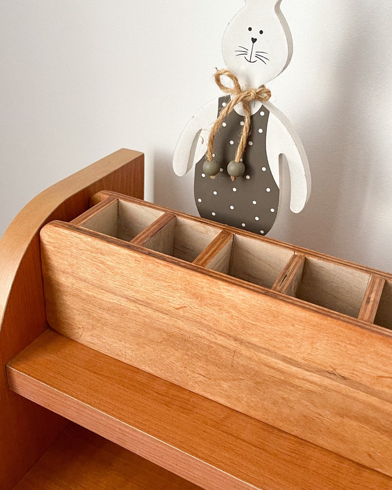 May include: A wooden desk organizer with a shelf and a compartment with multiple slots. The organizer is made of light brown wood and has a rounded top. A white wooden bunny figurine with a brown polka dot dress and a brown bow is standing on top of the organizer.