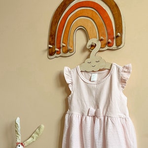 May include: A wooden rainbow-shaped wall hanger with five hooks. The rainbow is painted in shades of brown, orange, and red. A wooden cloud-shaped hanger with a smiling face is hanging from the rainbow.