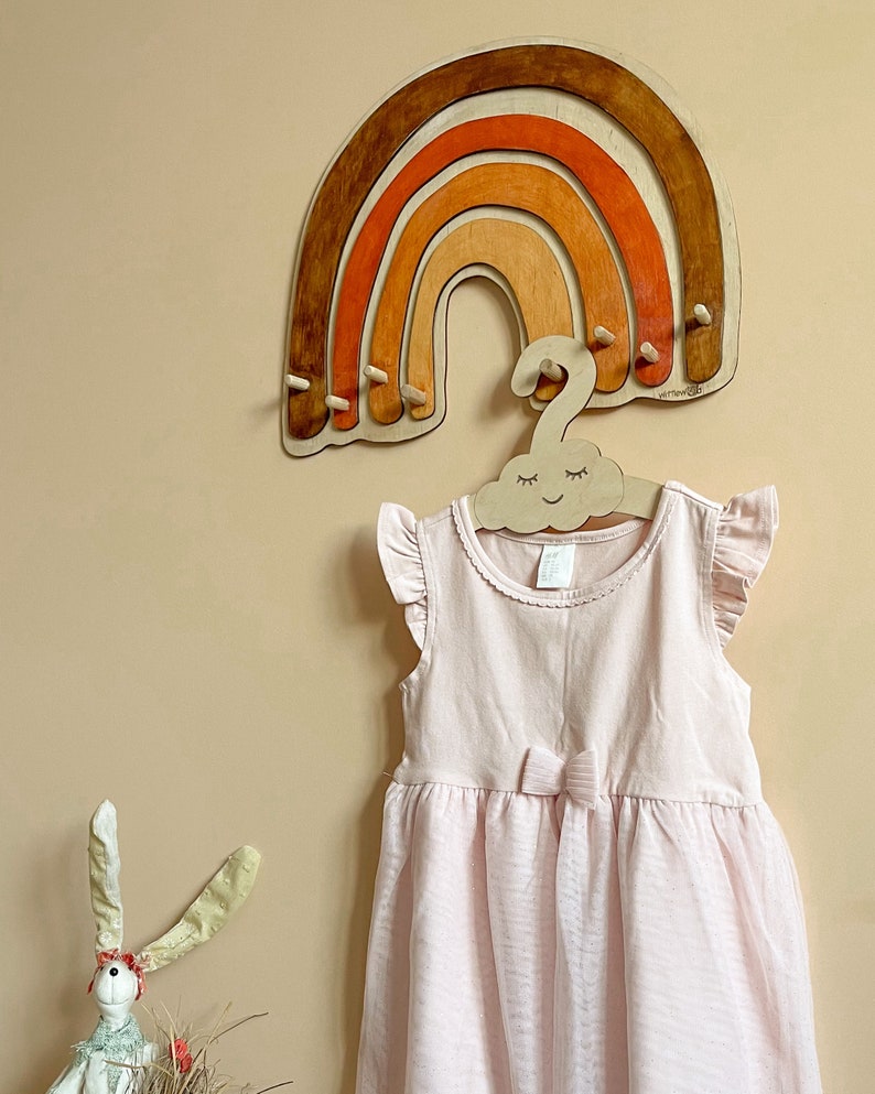 May include: A wooden rainbow-shaped wall hanger with five hooks. The rainbow is painted in shades of brown, orange, and red. A wooden cloud-shaped hanger with a smiling face is hanging from the rainbow.