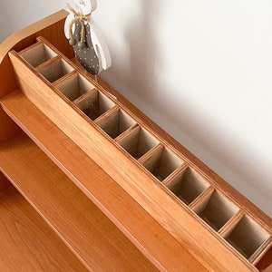 May include: A wooden organizer with multiple compartments, perfect for storing small items. The organizer is made of light brown wood and has a natural finish. It is sitting on a shelf made of the same wood.