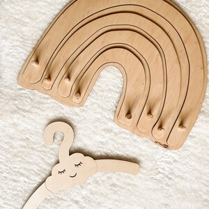 May include: Wooden rainbow-shaped hanger with five tiers and a cloud-shaped hanger with a smiling face and closed eyes. The rainbow hanger has the text "wittlewood" on the bottom.