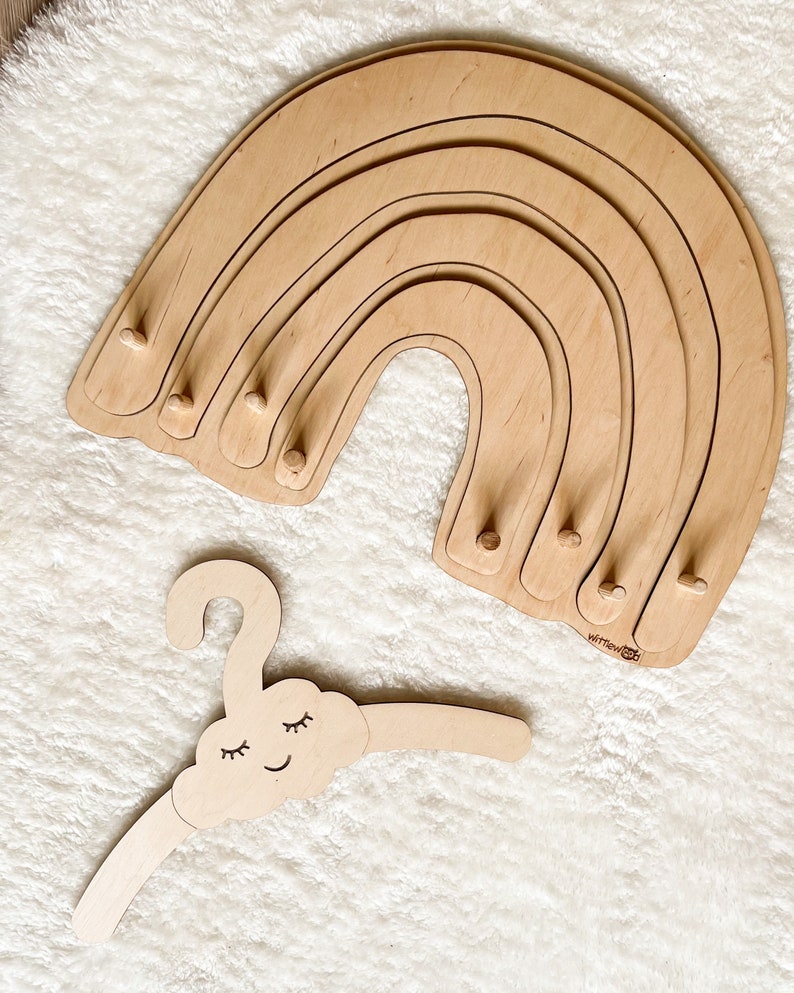 May include: Wooden rainbow-shaped hanger with five tiers and a cloud-shaped hanger with a smiling face and closed eyes. The rainbow hanger has the text "wittlewood" on the bottom.
