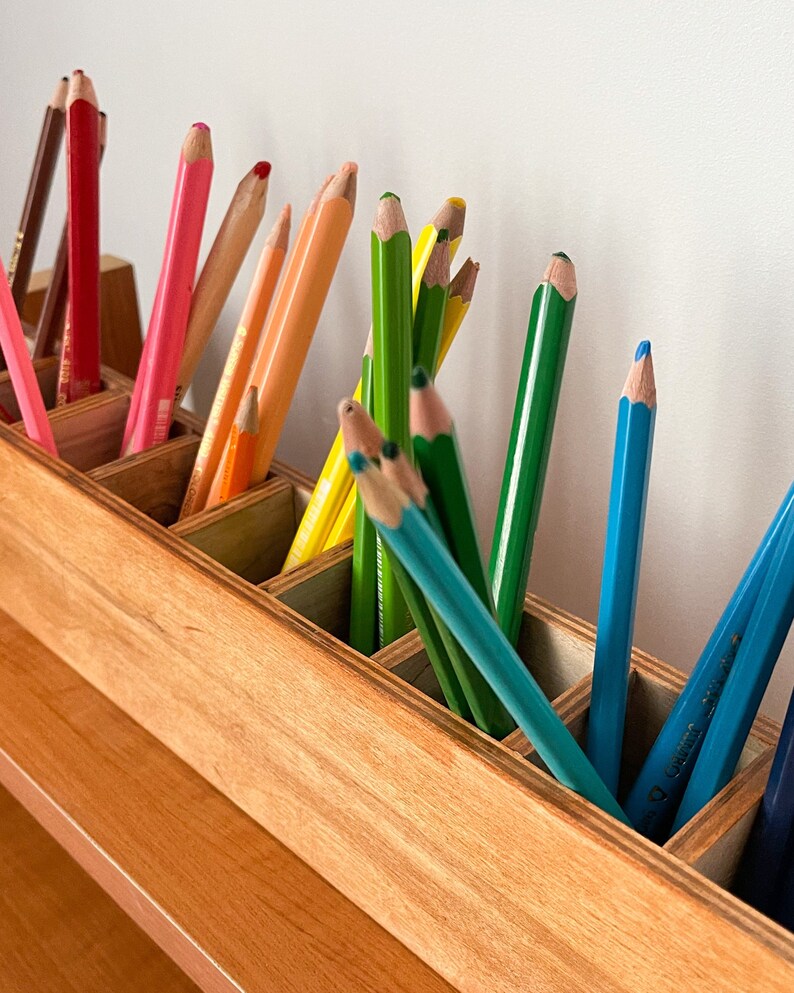 May include: A wooden pencil holder with multiple compartments, filled with colored pencils in shades of red, orange, yellow, green, blue, and brown.