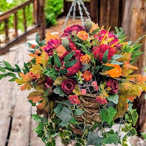 May include: A hanging basket filled with artificial autumn flowers and foliage, including deep red roses, orange blossoms, maple leaves, green ivy, and poppy seed pods. The arrangement is contained in a woven brown basket with a metal chain hanger.