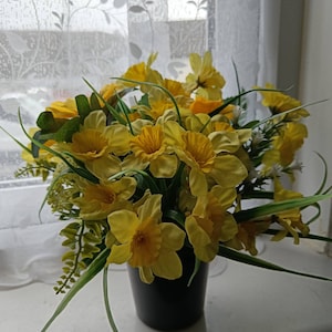 Puede incluir: Un ramo de narcisos amarillos en un jarrón negro. Las flores están dispuestas en un racimo y tienen hojas verdes.