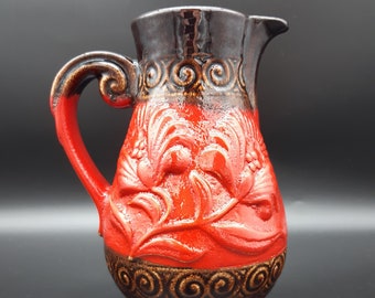 May include: A red and brown ceramic pitcher with a floral design. The pitcher has a wide base and a narrow neck with a spout. The handle is curved and has a decorative design.