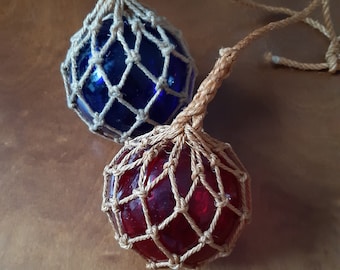 May include: Two glass fishing floats, one blue and one red, are wrapped in a natural-colored rope net. The floats are hanging from a rope.