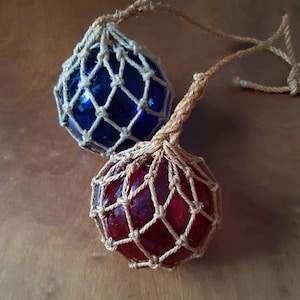 Op de afbeelding: Twee glazen visboeien, een blauwe en een rode, zijn gewikkeld in een natuurlijk gekleurd touwnet. De boeien hangen aan een touw.