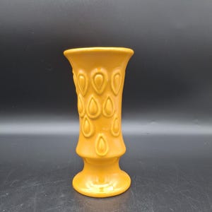 May include: A yellow ceramic vase with a textured design of teardrop shapes. The vase has a wide mouth and a narrow base.