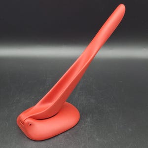 May include: A red, modern-style spoon rest with a curved handle and a wide, oval base. The spoon rest is made of a solid, matte material and is designed to hold a spoon or other utensil. The overall design is sleek and minimalist.