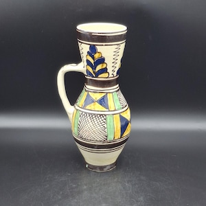 May include: A ceramic jug with a cream base and handle. It features geometric patterns in yellow, blue, and green, with a black and white grid design. The top section has a brown band with blue and yellow leaf-like designs.