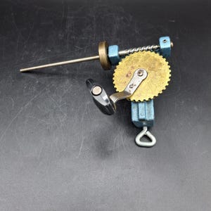 Könnte beinhalten: Eine Vintage-Handbohrmaschine mit einem Messingzahnrad, einem silbernen Bohrer und einem schwarzen Griff. Die Bohrmaschine hat blaue Metallkomponenten und eine silberne Metallschlaufe an der Basis. Das Werkzeug ist vor einem dunklen Hintergrund platziert.