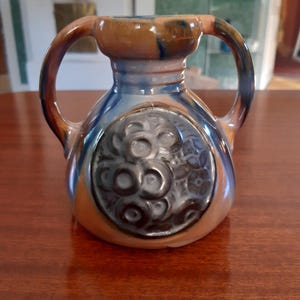 Könnte beinhalten: Eine kleine, braune, blaue und weiße Keramikvase mit zwei Henkeln. Die Vase hat ein erhabenes, kreisförmiges Design mit einem floralen Muster.