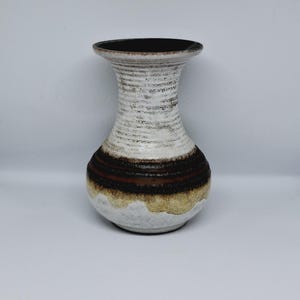 Peut inclure: Un vase en céramique avec un col évasé et une base bulbeuse. Le col présente une surface texturée et nervurée blanc cassé avec des taches brun foncé. La partie centrale possède une large bande brun foncé, tandis que la base est beige moucheté.