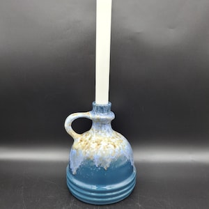 May include: A ceramic candle holder with a handle, in shades of blue and tan, holds a tall white candle. The holder has a rounded body with horizontal ridges and a flared top. The candle is centered in the holder.