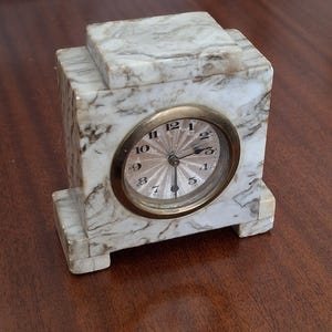 May include: A marble-patterned, rectangular clock with a round clock face. The clock face has a gold-colored bezel and a light-colored face with black numerals. The clock is set on a dark brown surface.