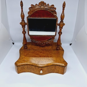 May include: An antique wooden vanity mirror with a small drawer. The mirror is rectangular and framed by a decorative wooden border with a red velvet backing. The base and frame are made of a light brown wood.
