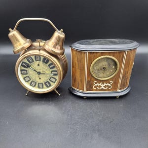 May include: Two antique clocks, one is a brass alarm clock with Roman numeral markings and the other is a wooden clock with a black face and Arabic numeral markings.