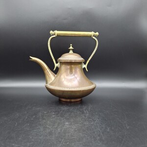 Könnte beinhalten: Eine Vintage-Teekanne aus Kupfer mit Messinggriff und Ausguss. Die Teekanne hat einen abgerundeten Körper mit dekorativem Perlenbesatz und einem kleinen Knauf auf dem Deckel. Der Griff ist geschwungen und der Ausguss schlank. Die Teekanne wird wahrscheinlich zum Brühen von Tee verwendet.