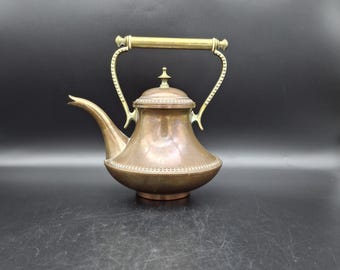 Pode incluir: Uma chaleira de cobre vintage com uma alça e bico de latão. A chaleira tem um corpo arredondado com uma borda decorativa de contas e um pequeno botão na tampa. A alça curva-se graciosamente e o bico é fino. A chaleira é provavelmente usada para preparar chá.