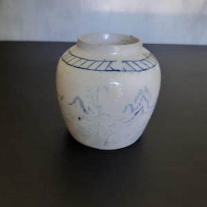 May include: A small white ceramic jar with blue and white painted designs. The jar has a rounded body and a narrow opening. The design features a landscape scene with mountains, trees, and a river.