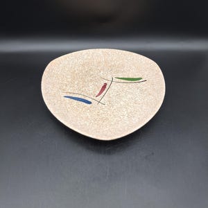 May include: A beige, mid-century modern style plate with an asymmetrical shape. The plate features abstract designs in blue, red, and green. The plate is made of ceramic and is suitable for serving appetizers or small dishes.