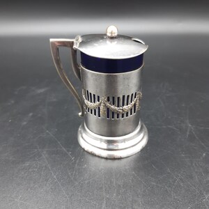 Puede incluir: Un dispensador de azúcar de plata con un revestimiento de vidrio azul y una tapa decorativa. El dispensador tiene un asa y un diseño decorativo alrededor del medio.