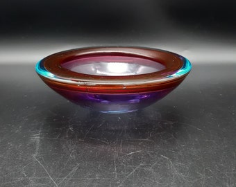 Könnte beinhalten: Eine kleine, runde Glasschüssel mit einem geschichteten Design. Die Schüssel besteht aus klarem Glas mit einem Farbspiel aus Lila, Rot und Blau.