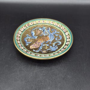 May include: An oval decorative dish with a gold-coloured rim and a textured, gold-coloured interior. The dish features a detailed design of a bird and floral elements in blue, pink, and green. The rim is decorated with small red and white dots.